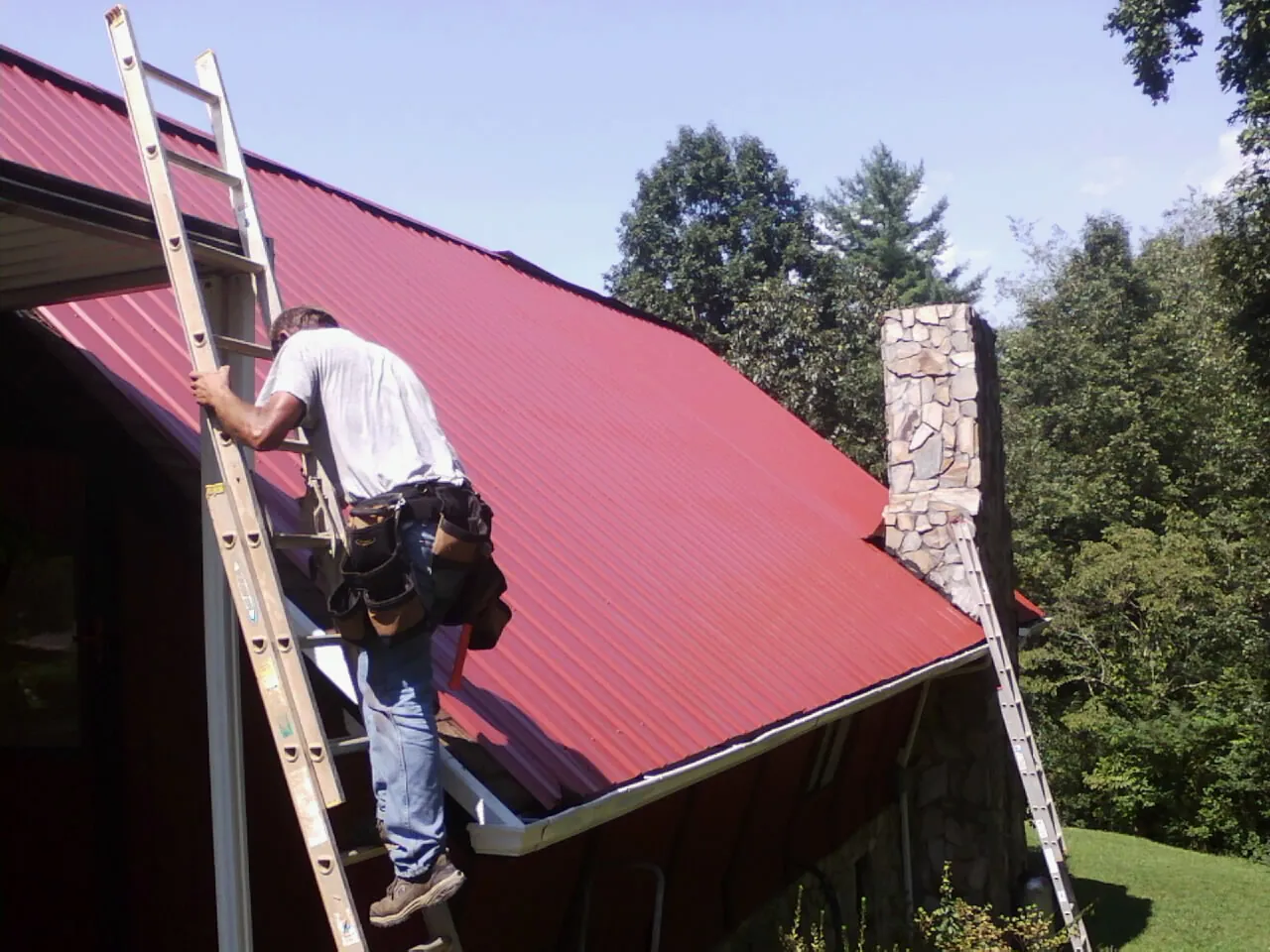 Roofer on a ladder beside a red metal roof and stone chimney — Soffit & Fascia Repair by Roofing Brothers in Gervais